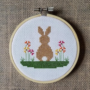 May include: A brown bunny rabbit cross-stitch pattern with yellow, orange, and pink flowers on a green patch of grass. The cross-stitch is framed in a wooden hoop.