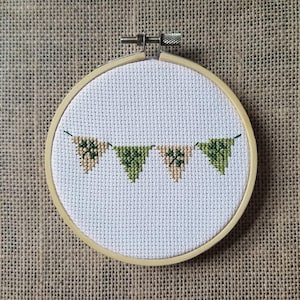 May include: A cross-stitch embroidery hoop with a banner of green and beige triangle flags. The flags are stitched on white fabric and framed by a wooden hoop. The background is a textured, beige fabric.
