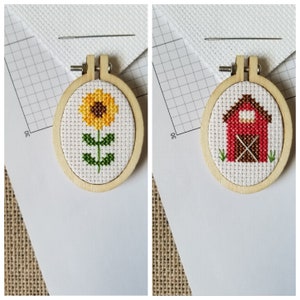 May include: Two small, oval wooden embroidery hoops each hold a cross-stitch design. One features a yellow sunflower with a green stem and leaves. The other depicts a red barn with a brown door and green grass. A needle and fabric are in the background.