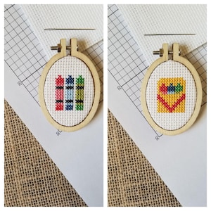 May include: Two small, round, wooden embroidery hoops with white fabric inside. The left hoop has a cross-stitch design of three crayons in red, pink, blue, and green. The right hoop has a cross-stitch design of a heart with crayons inside.