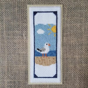 Könnte beinhalten: Ein Kreuzstich-Lesezeichen mit einer Küstenlandschaft. Das Design zeigt eine Möwe, blaues Meer, gelbe Sonne und eine weiße Wolke. Das Lesezeichen hat einen marineblauen Rahmen und einen beigen Hintergrund.