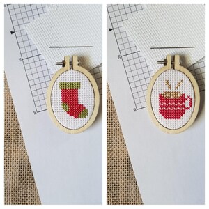 May include: Two small, oval wooden embroidery hoops each feature a cross-stitched design. One depicts a red and green Christmas stocking, the other a red mug with steam. A needle and white fabric are visible.