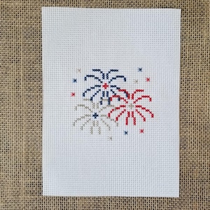 May include: Cross-stitch design featuring three fireworks in red, white, and blue, with star accents. The design is on a white fabric background, perfect for patriotic crafts or home decor.