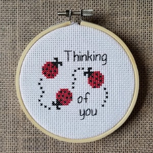 Può includere: Un cerchio da ricamo bianco con un disegno a punto croce di tre coccinelle rosse e il testo "Thinking of you".