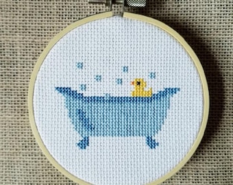 May include: A white cross-stitch embroidery hoop with a blue bathtub and a yellow rubber ducky inside. The ducky is floating in the bathtub with blue bubbles around it.