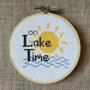 Puede incluir: Un aro de bordado de punto de cruz blanco con un sol amarillo, olas azules y el texto "on Lake Time".