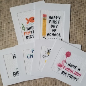May include: A collection of handmade cross-stitch cards with white frames. Designs include a goldfish, a pencil with "Happy First Day of School", a bee, and a flamingo with "Have a Fabulous Birthday". The cards are on a textured surface.