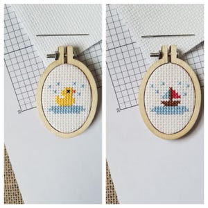 May include: Two small wooden embroidery hoops with cross-stitch designs. The left hoop features a yellow rubber duck in a blue bathtub with blue raindrops. The right hoop features a red and white sailboat on blue water with white raindrops.
