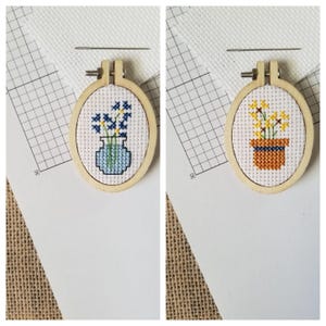 Puede incluir: Dos pequeños bastidores de bordado de punto de cruz ovalados. Uno presenta flores azules en un jarrón azul, el otro flores amarillas en una maceta marrón. Ambos están en aros de madera, con una aguja encima.