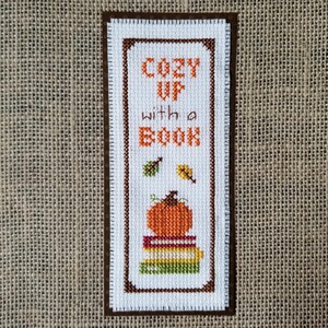 Puede incluir: Marcapáginas bordado con borde marrón y el texto "COZY UP with a BOOK". El marcapáginas presenta una calabaza sobre una pila de libros, con hojas. La combinación de colores incluye naranja, verde y marrón.