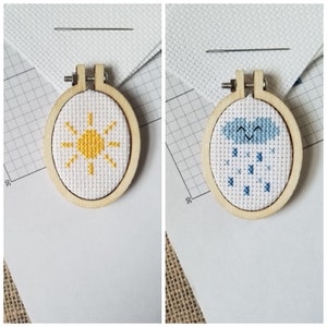 Pode incluir: Dois pequenos aros de bordado de madeira com tecido branco no interior. Um aro apresenta um sol amarelo com raios, o outro uma nuvem azul com gotas de chuva.
