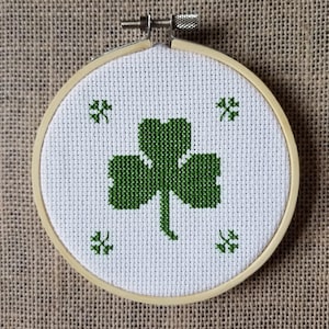 Puede incluir: Un aro de bordado blanco con un diseño de trébol verde. El trébol está bordado con hilo verde sobre un fondo de tela blanca.