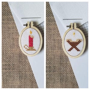 May include: Two small, wooden embroidery hoops each feature a cross-stitch design. One depicts a red candle with a yellow flame, and the other shows a manger with a star. Both are on white fabric with a needle.