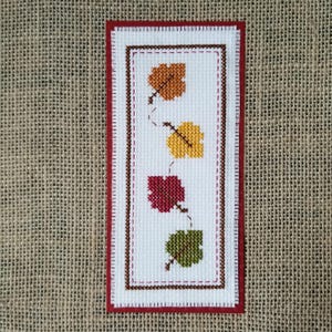 May include: A rectangular cross-stitch design featuring four autumn leaves in shades of orange, yellow, red, and green. The leaves are stitched on a white background, framed by a red border and a brown stitched outline. The background is a burlap texture.