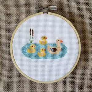Puede incluir: Bordado de punto de cruz de un pato y tres patitos nadando en un estanque azul, con juncos a la izquierda. El bordado está en un aro de madera redondo, con un fondo de tela beige.