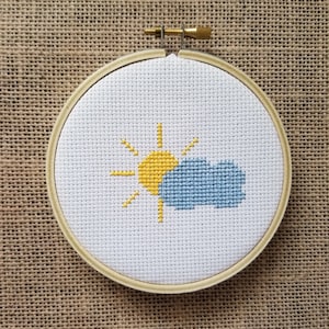 Op de afbeelding: Een witte borduurring met een kruissteekontwerp van een gele zon met stralen en een blauwe wolk.