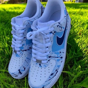 Puede incluir: Zapatillas blancas con diseño de pintura salpicada azul y marino. Los zapatos presentan cordones blancos, un logotipo de Nike azul y una suela blanca. Las zapatillas están sobre una superficie de hierba verde.