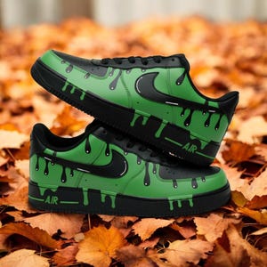May include: Black and green trainers with a drip design. The trainers have a black swoosh and the word "AIR" on the side. The shoes are set against a backdrop of autumn leaves.