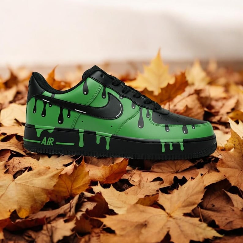 May include: A custom green and black trainer with a dripping paint design. The shoe features a black swoosh, laces, and sole, with the word "AIR" printed on the side. The trainer is set against a backdrop of autumn leaves.