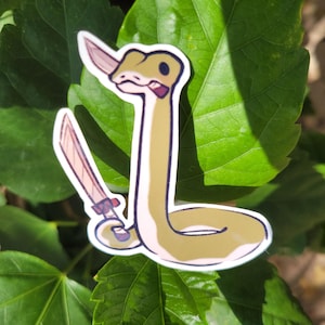 May include: A sticker of a cartoon snake holding a knife. The snake is light green and tan with a white border. The knife is pink and tan. The sticker is placed on a background of green leaves.