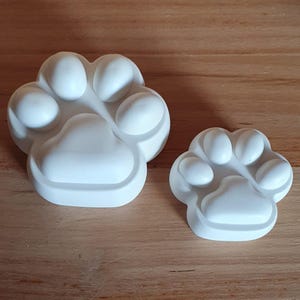 May include: Two white paw print shaped objects. One is larger than the other. The paw prints have a rounded shape with five toes and a larger pad. The objects are sitting on a wooden surface.