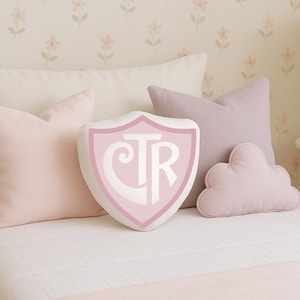 May include: A bed with several pillows in shades of pink and lavender. A shield-shaped pillow with the letters "CTR" is centered. The bed is made with a white comforter and a pink bed runner. The background features a floral wallpaper.