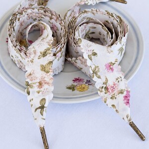 May include: Two pairs of white floral shoelaces with scalloped edges and brass aglets are coiled on a floral patterned plate. The laces feature pink and green floral designs. The word "White" is at the bottom of the image.