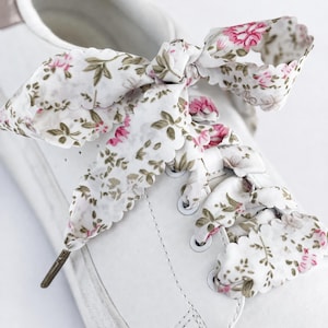 May include: White trainers with floral print laces. The laces display a white background adorned with pink flowers and green leaves. The laces are tied in a bow, enhancing the aesthetic appeal of the footwear.