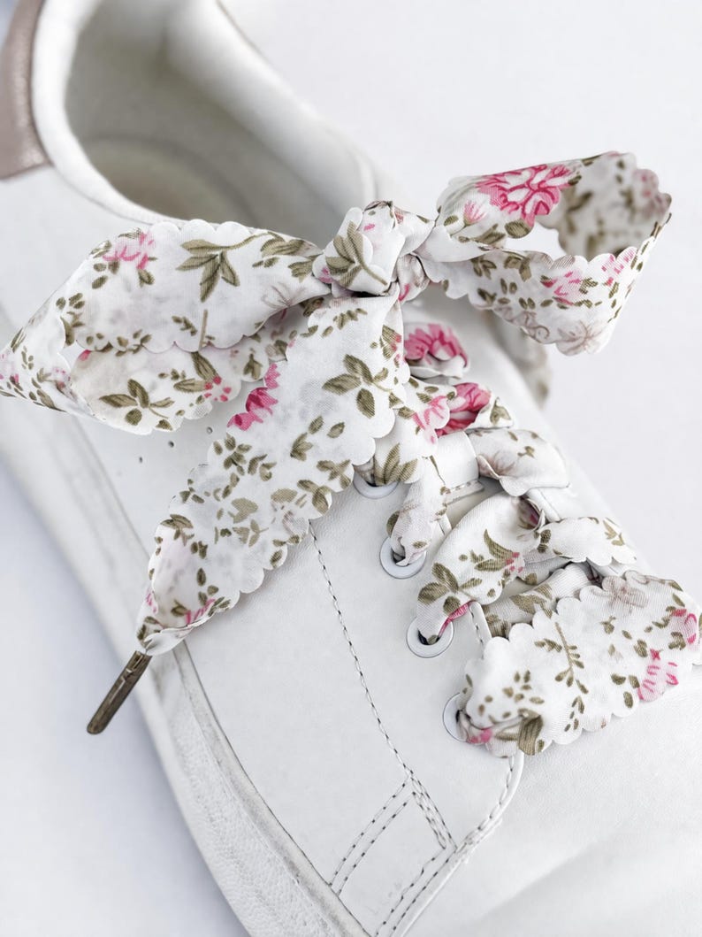 May include: White trainers with floral print laces. The laces display a white background adorned with pink flowers and green leaves. The laces are tied in a bow, enhancing the aesthetic appeal of the footwear.