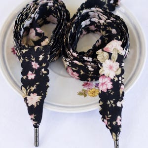May include: Black floral patterned shoelaces with scalloped edges and metal aglets. The laces are arranged in a figure-eight pattern on a white plate with floral accents. The word "BLACK" is printed at the bottom.
