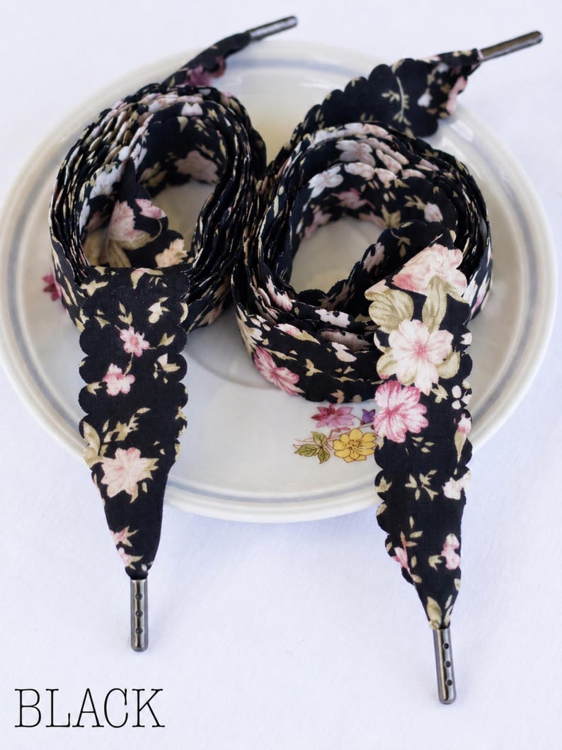 May include: Black floral patterned shoelaces with scalloped edges and metal aglets. The laces are arranged in a figure-eight pattern on a white plate with floral accents. The word "BLACK" is printed at the bottom.