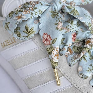 May include: A close-up of a white Adidas Gazelle trainer with a light blue floral patterned shoelace bow. The shoelace has a gold aglet. The word "Gazelle" is printed in gold on the side of the shoe. The trainer has white stripes.