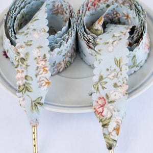 May include: Two spools of light blue floral ribbon with scalloped edges and gold aglets. The ribbon features a delicate pattern of pink, white, and green flowers. The word "BLUE" is printed at the bottom of the image.