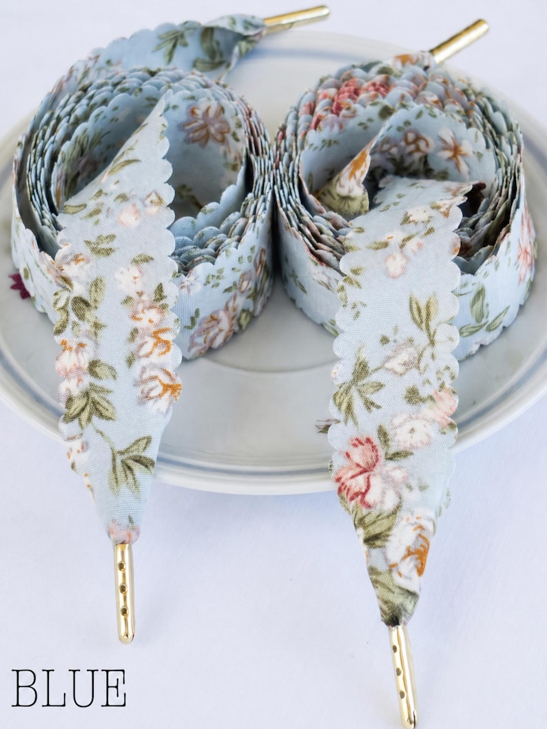 May include: Two spools of light blue floral ribbon with scalloped edges and gold aglets. The ribbon features a delicate pattern of pink, white, and green flowers. The word "BLUE" is printed at the bottom of the image.