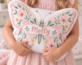 May include: A butterfly-shaped throw pillow featuring a white background with a watercolor-style floral pattern in shades of pink, green, and yellow. The center of the butterfly displays the name Molly in a pink script font on a white oval.