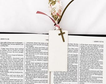 May include: A rectangular white bookmark with stitched edges, featuring a bronze-colored cross charm attached to the top. A fabric ribbon with a floral pattern in pink, green, and white is tied to the bookmark, resting on an open printed book.