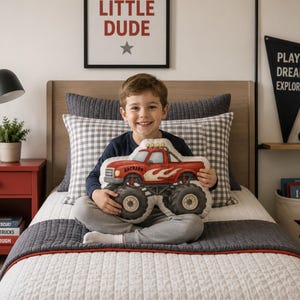 May include: A plush throw pillow shaped like a red monster truck with large black tires, white flame decals on the side, and the name Jackson printed in black script on the door. The truck features yellow headlights and a roll bar on the roof.
