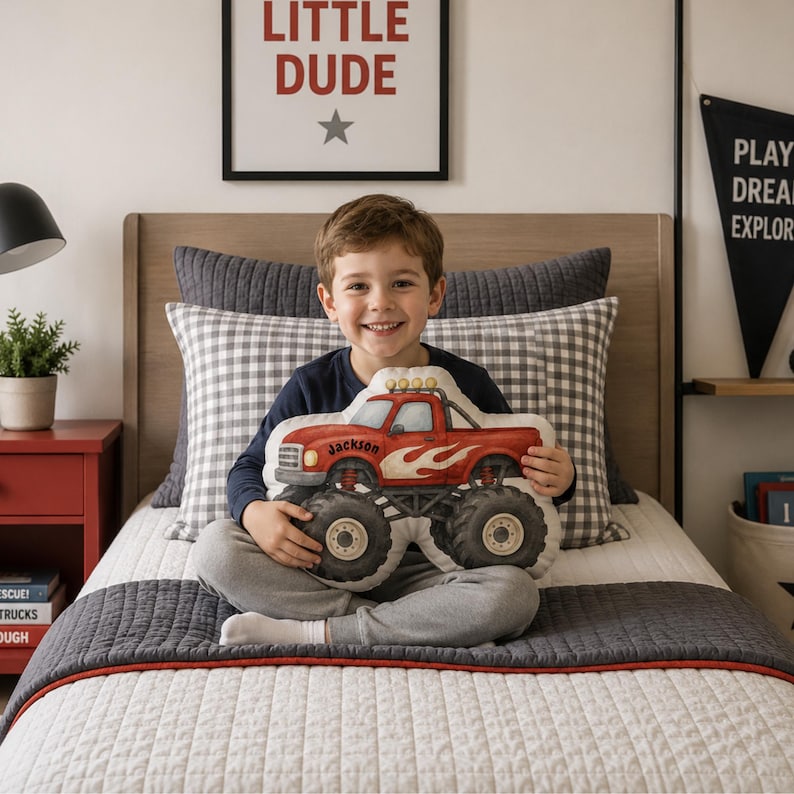 May include: A plush throw pillow shaped like a red monster truck with large black tires, white flame decals on the side, and the name Jackson printed in black script on the door. The truck features yellow headlights and a roll bar on the roof.