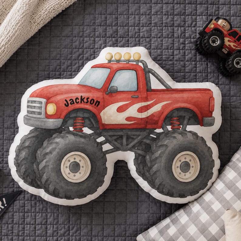 May include: A shaped throw pillow designed as a red monster truck with large black tires, white flame graphics on the side, and the name Jackson printed in black script on the hood. The truck features a roll bar with four yellow lights on top.