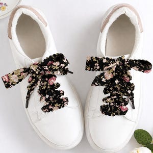 May include: White trainers with floral patterned laces. The trainers have a white leather upper, a rounded toe, and a light brown trim around the ankle. The laces are black with a pattern of pink and cream-colored flowers.