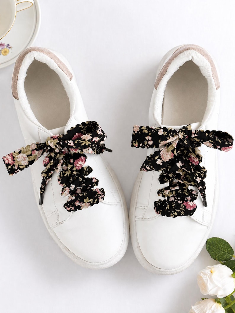 May include: White trainers with floral patterned laces. The trainers have a white leather upper, a rounded toe, and a light brown trim around the ankle. The laces are black with a pattern of pink and cream-colored flowers.