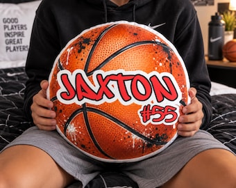 May include: A round basketball-themed throw pillow featuring a textured orange surface with black lines and black paint splatter accents. The pillow displays the name JAXTON in bold red letters with a white outline, accompanied by the number #55 in red.