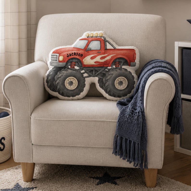 May include: A shaped throw pillow designed as a red monster truck with large black tires, flame graphics on the side, and the name Jackson printed in black script on the door. The pillow features a roll bar with four circular lights on top.