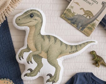 May include: A shaped throw pillow featuring a printed illustration of a green dinosaur with dark stripes and the name ROWAN printed on its tail. The dinosaur has a light tan underbelly, dark claws, and a white border around the entire shape.