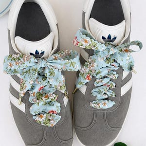 May include: Grey suede trainers with white detailing and floral ribbon laces. The Adidas logo is visible on the tongue and side. The light blue laces have a floral pattern in red, white, and green. The trainers are a stylish footwear option.
