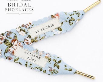 May include: Light blue shoelaces with a floral pattern, scalloped edges, and gold-toned metal aglets. One lace features a white rectangular patch with the text 11.12.2026, and the other features a patch with the text ALLISON + ZURI.
