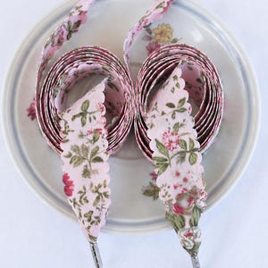 May include: Pink floral shoelaces with scalloped edges and silver aglets are coiled on a floral patterned plate. The shoelaces have a pink background with a repeating pattern of red and green flowers and leaves.