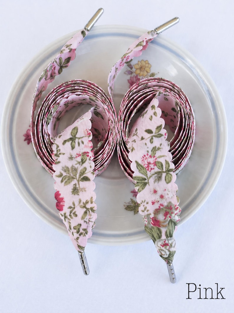 May include: Pink floral shoelaces with scalloped edges and silver aglets are coiled on a floral patterned plate. The shoelaces have a pink background with a repeating pattern of red and green flowers and leaves.
