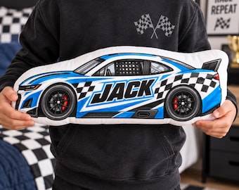 May include: A plush pillow shaped like a race car, featuring a blue and white color scheme with black accents. The side of the car displays the name JACK in bold black letters, along with a checkered flag pattern near the rear wheel.