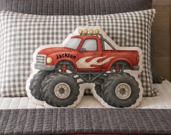 May include: A shaped throw pillow featuring a red monster truck with large black tires, a roll bar with four yellow lights, and white flame graphics on the side. The name Jackson is printed in black script on the side of the truck cab.